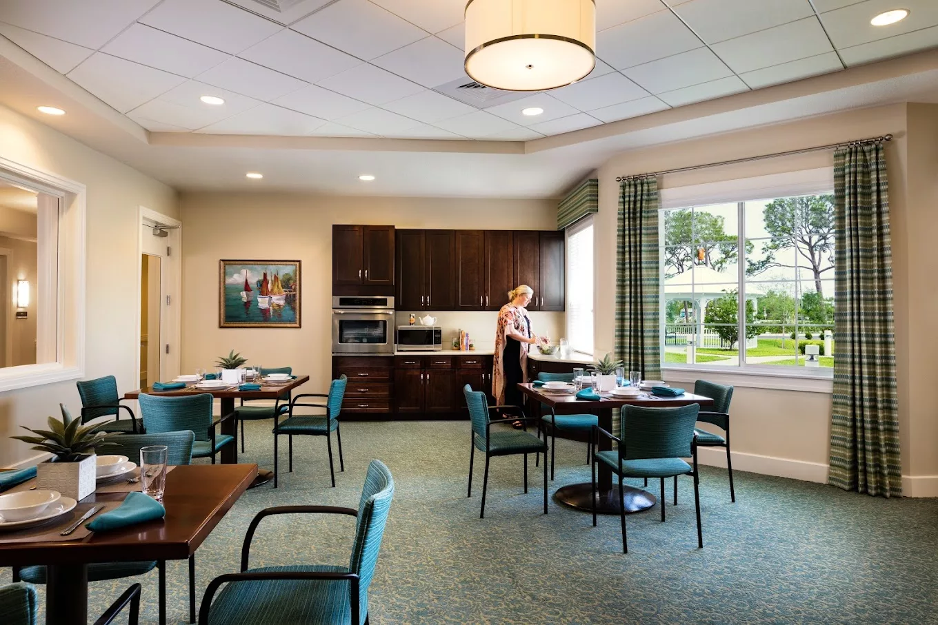 A modern dining room with a kitchen area, featuring tables and chairs, where a woman is setting up the space.