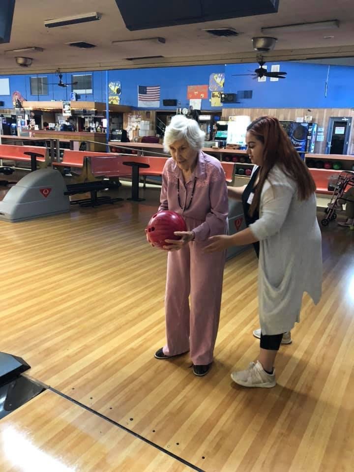 An elderly woman receives assistance while bowling at an indoor alley.