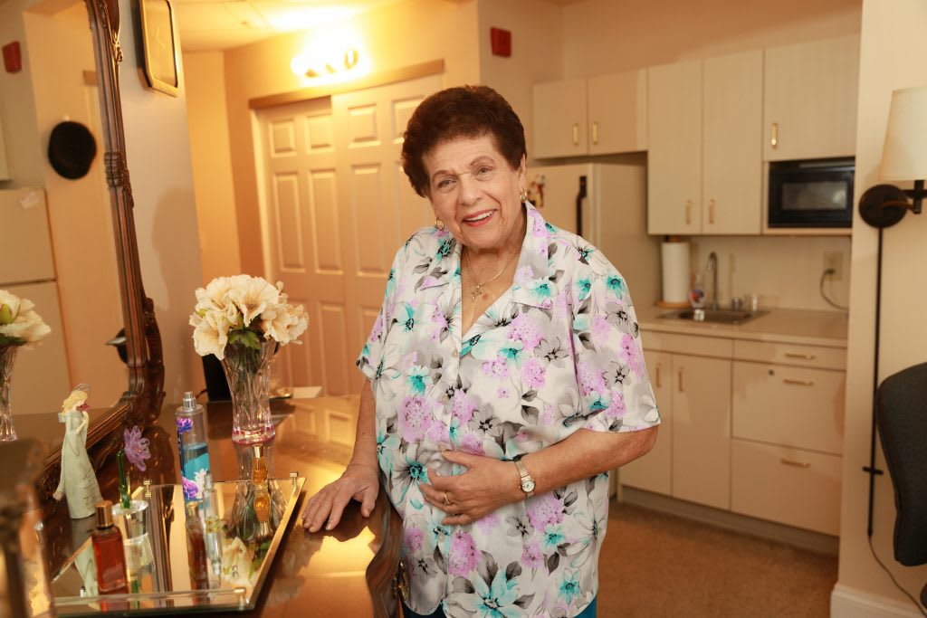 An elderly person smiling in a kitchen area, wearing a floral shirt with flowers on the counter.