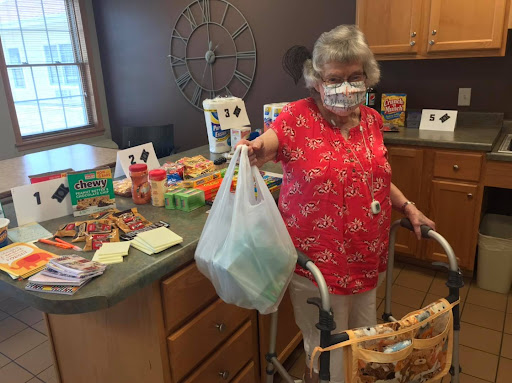 An elderly woman in a red blouse is holding a bag of groceries while using a walker in her kitchen.