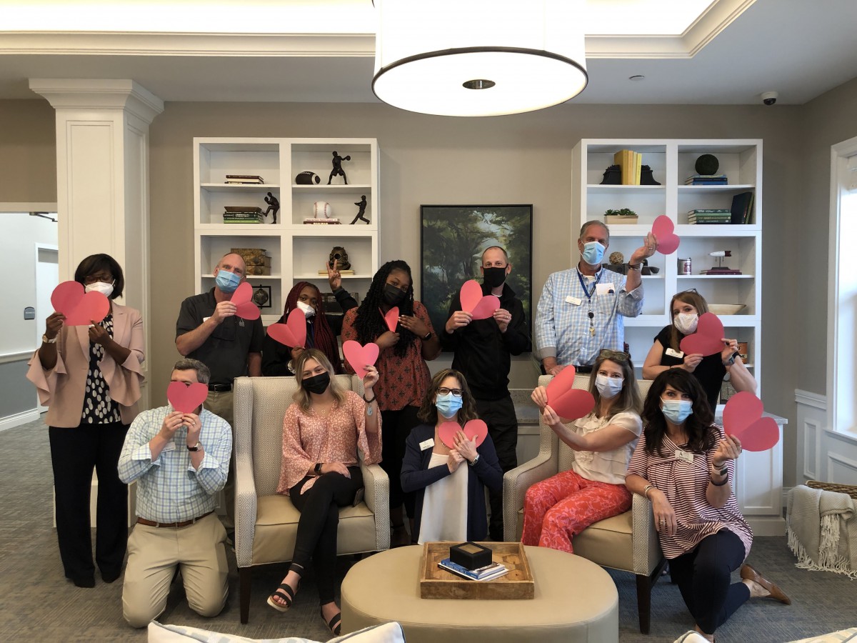 A group of people posing indoors, many wearing masks and holding pink paper hearts.