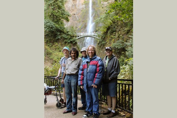 A group of people standing in front of a waterfall and a bridge, enjoying the scenic outdoor setting.