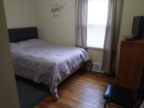 A cozy bedroom with a neatly made bed, wooden flooring, and natural light streaming through the window.