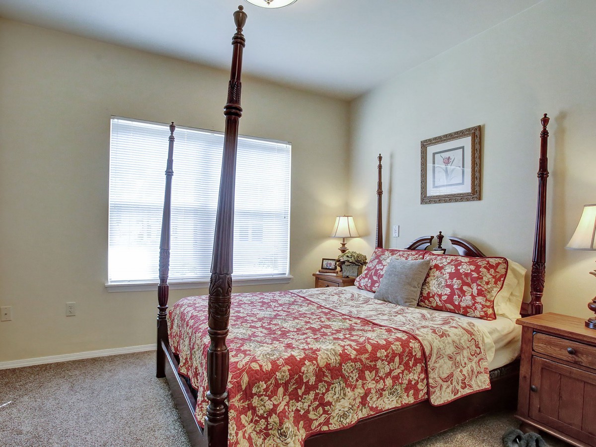 A cozy bedroom featuring a four-poster bed with floral bedding, a bedside table with a lamp, and a window with blinds.