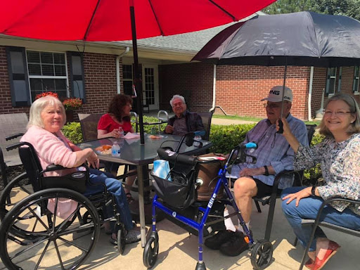 A group of senior citizens enjoying a sunny day outside, gathered around a table under a large red umbrella.