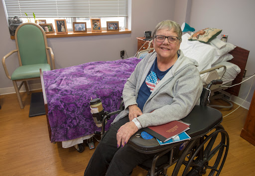 A smiling individual seated in a wheelchair, surrounded by personal belongings in a cozy room.