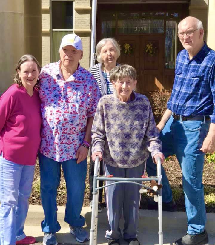 A group of five seniors standing together outdoors, with one using a walker.
