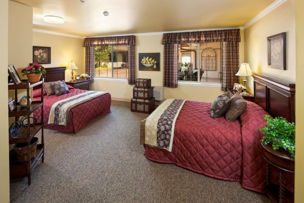 A spacious hotel room featuring two double beds with decorative blankets, bedside lamps, and large windows providing natural light.