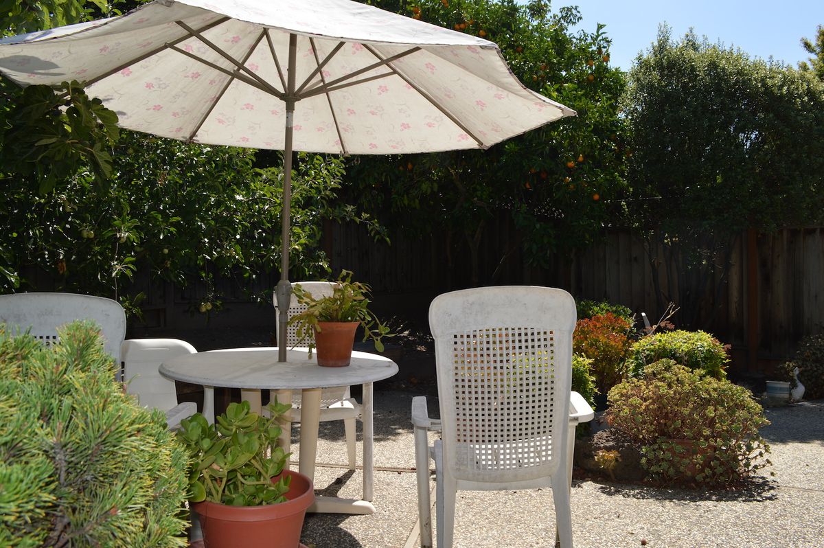 A sunny patio with a white table and chairs beneath a large umbrella, surrounded by potted plants.