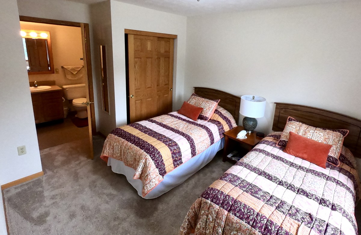 A cozy bedroom featuring two twin beds with quilted bedspreads, a shared nightstand with a lamp, and a doorway leading to a bathroom.