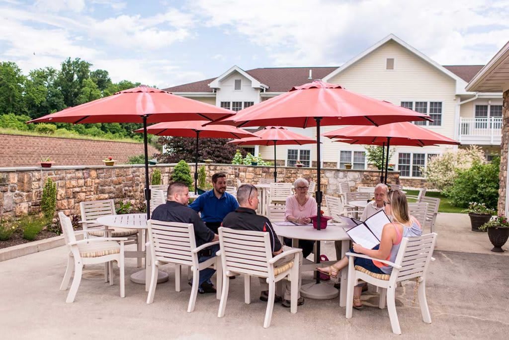 A group of people sitting at a table under red umbrellas, engaged in conversation on an outdoor patio.