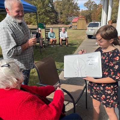 A young girl shows a drawing on a whiteboard to an elderly woman, with a man watching nearby during an outdoor gathering.
