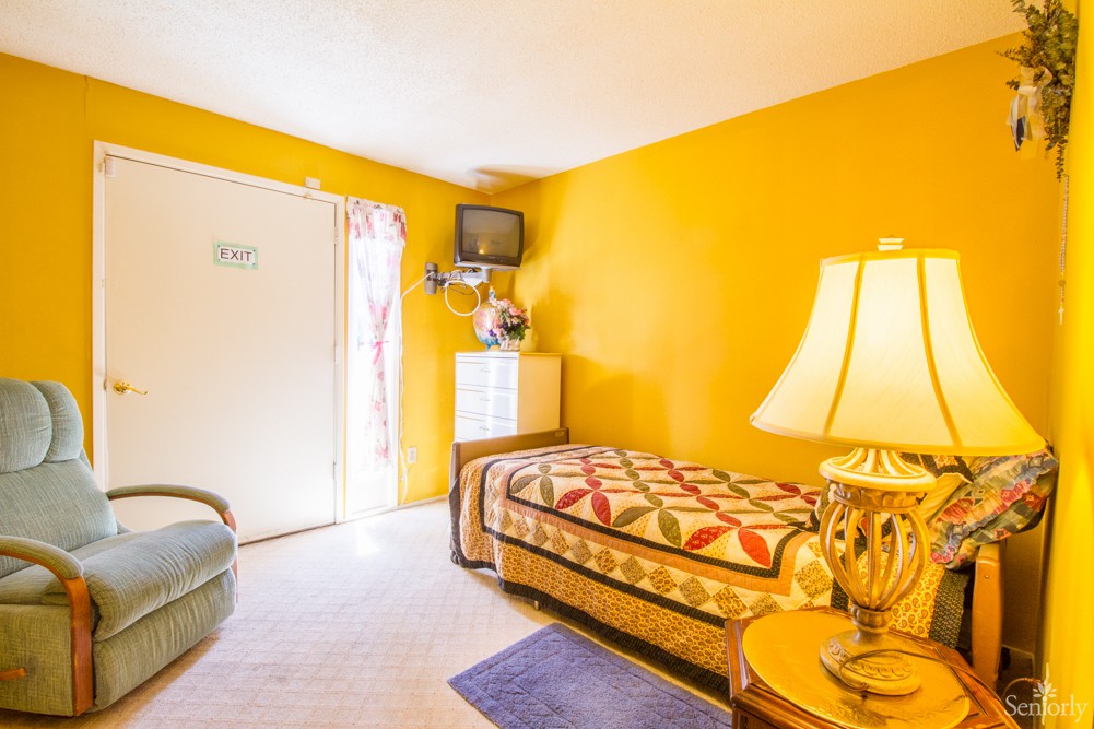 A cozy, brightly colored bedroom with a single bed, lamp, and mounted television.