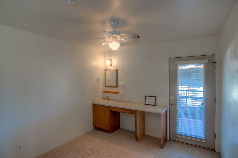 A minimalistic room with a small desk, mirror, ceiling fan, and carpeted floor.