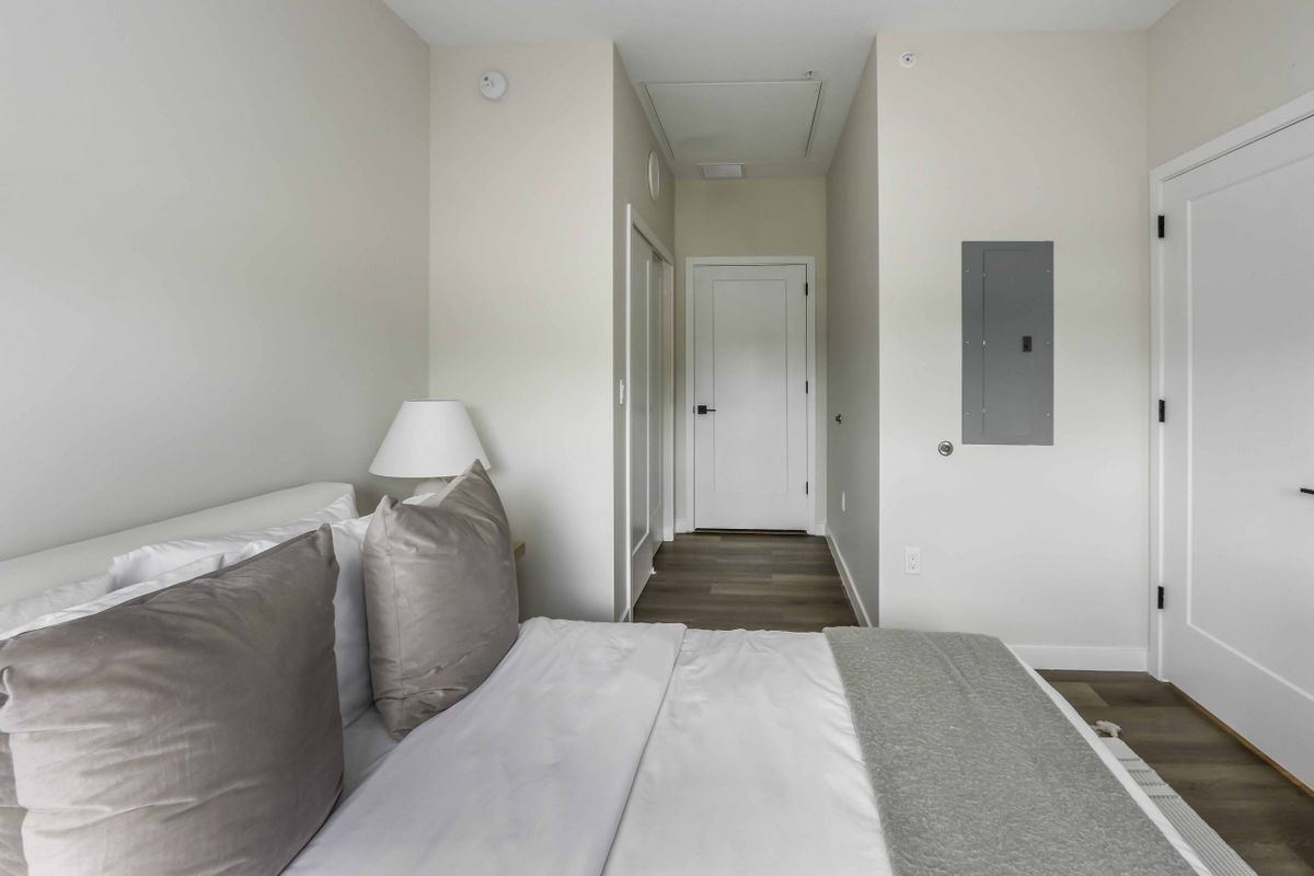 A minimalist bedroom featuring a neatly made bed with plush pillows and a lamp, leading to a white doorway.