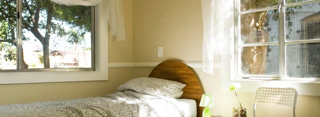 A cozy bedroom corner with a bed, pillow, and bedside lamp near a window with sheer curtains.