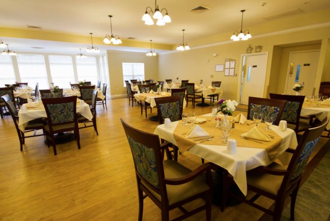 A warmly lit dining room with several set tables and chairs, featuring elegant chandeliers and a wooden floor.