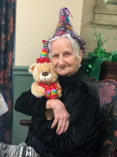 An elderly woman in a party hat joyfully holding a stuffed bear, celebrating her special day.