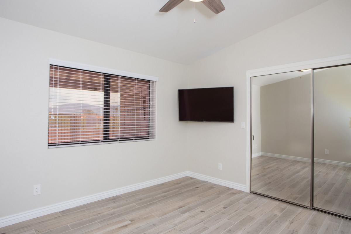 An empty room with wood flooring, featuring a mounted TV, window blinds, and a mirrored closet door.
