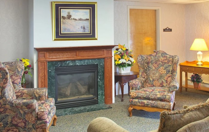 A cozy living room with a decorative fireplace, floral armchairs, and a landscape painting above the mantel.