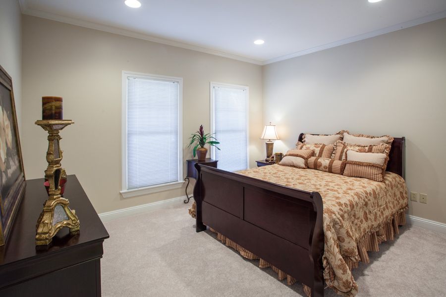 A cozy, well-lit bedroom featuring a wooden bed with decorative pillows, dual windows, and a bedside lamp.