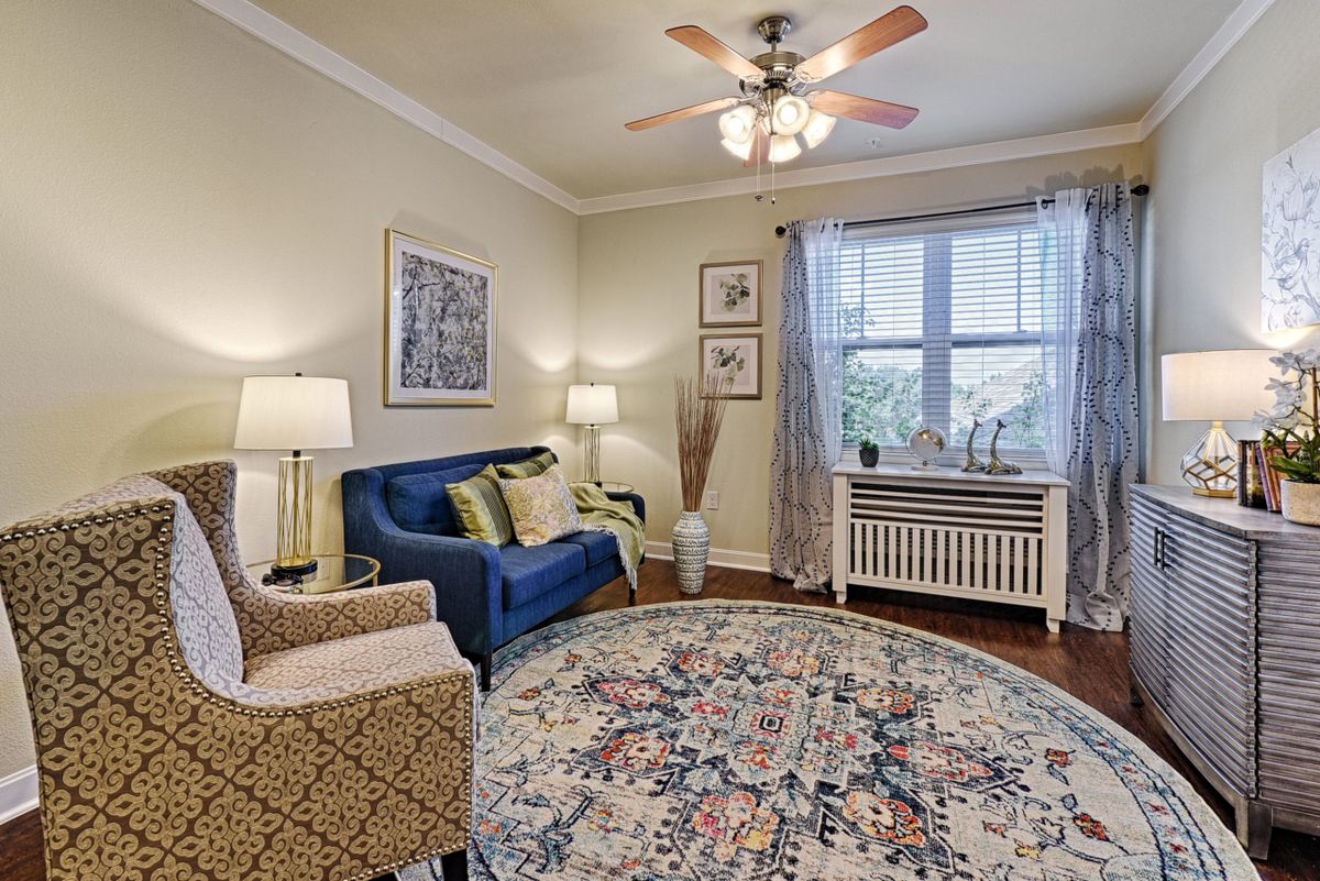 A cozy living room featuring a blue sofa, patterned armchair, round rug, and ample lighting from lamps and a ceiling fan.