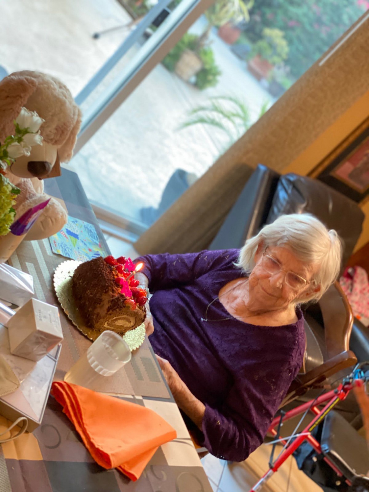An elderly woman sits at a table with a birthday cake and flowers, celebrating a special occasion.