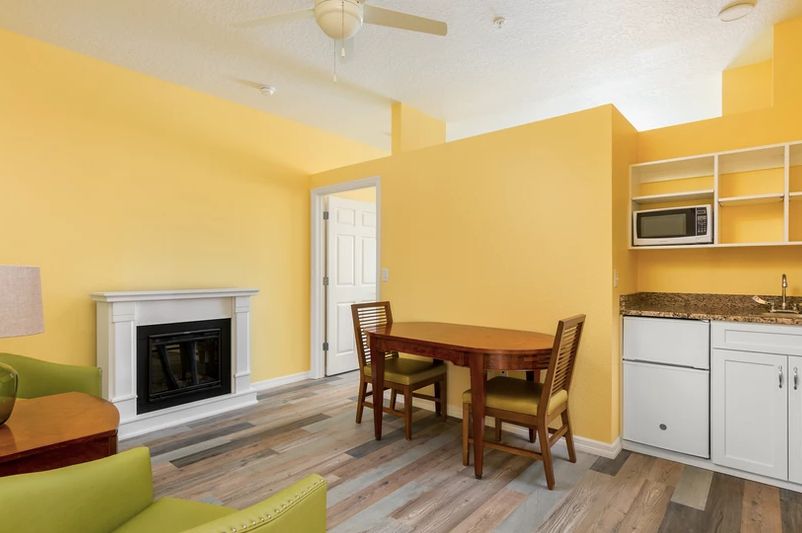 A cozy living room with yellow walls featuring a dining table and chairs, a fireplace, and an adjacent kitchen area.