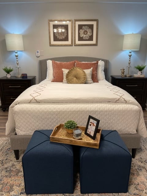 A cozy bedroom setup featuring a neatly made bed with decorative pillows, flanked by nightstands and lamps, complemented by a tray on ottomans.
