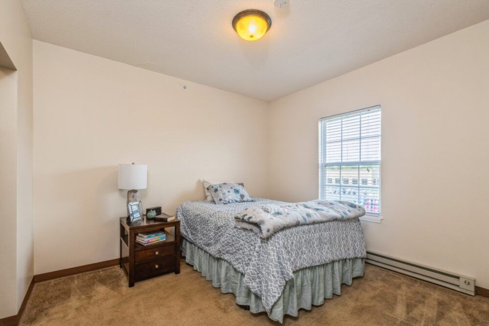 A cozy bedroom with a neatly made bed, nightstand, and a window letting in natural light.
