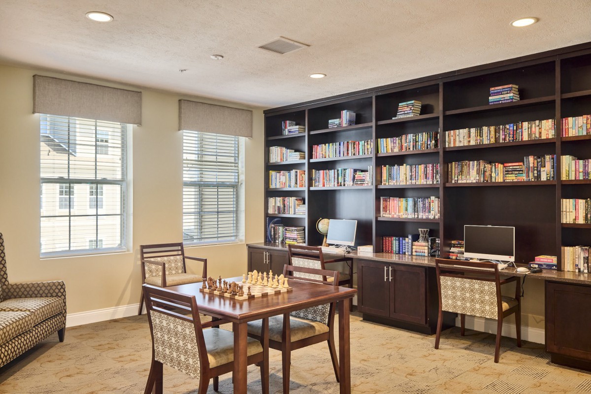 A cozy home office featuring bookshelves, a desk, and a chess set on the table by the window.