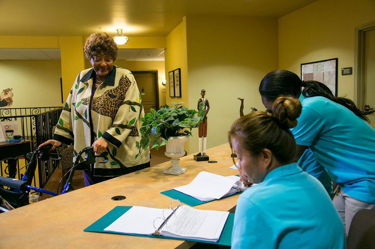 An elderly woman approaches the reception desk at a care facility, where staff are taking notes.