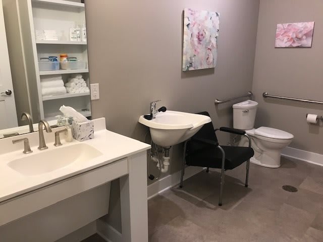 A tidy bathroom with a sink, toilet, and a chair next to shelves holding towels.