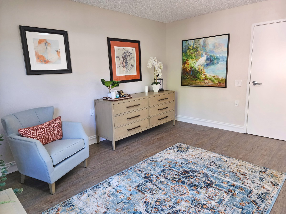 A cozy living room with a light blue chair, a wooden dresser adorned with plants and decorative items, and framed artwork on the walls.
