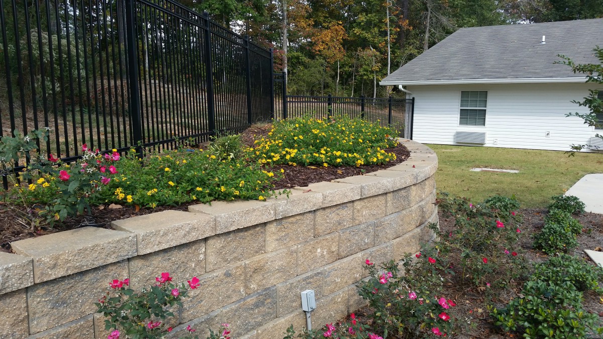 A landscaped garden featuring a stone retaining wall adorned with blooming flowers, bordered by a tall fence.