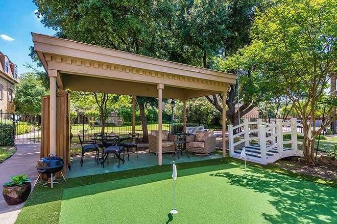 A cozy outdoor patio featuring a putting green and comfortable seating under a gazebo, surrounded by lush greenery.