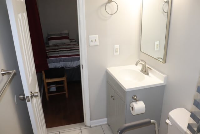 A view of a bathroom with a sink and mirror, leading into a bedroom.