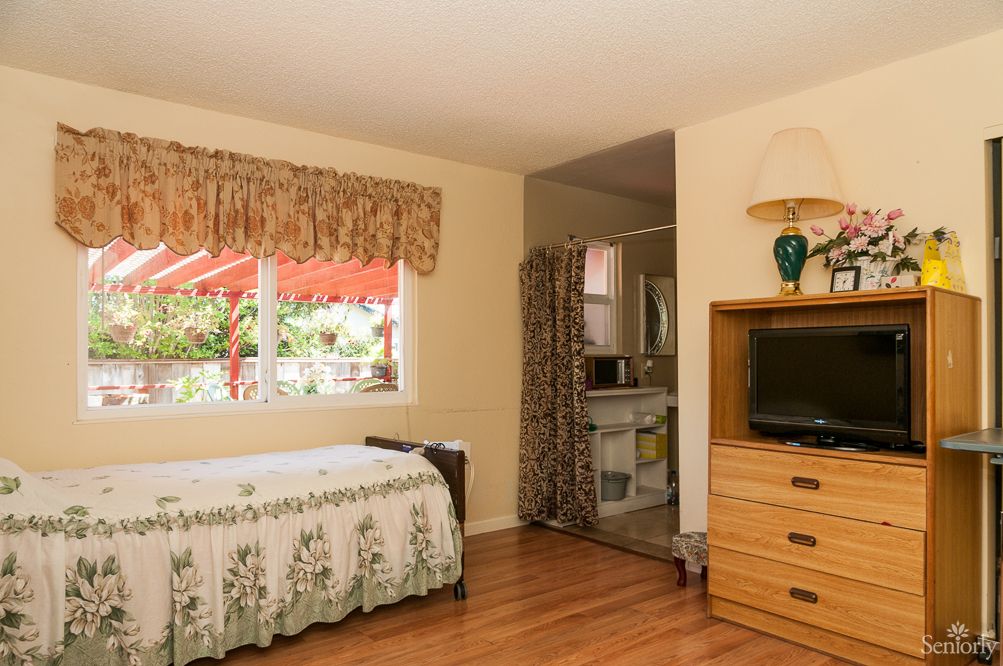 A cozy bedroom featuring a bed, TV, and dresser with a view of the outside through a window.