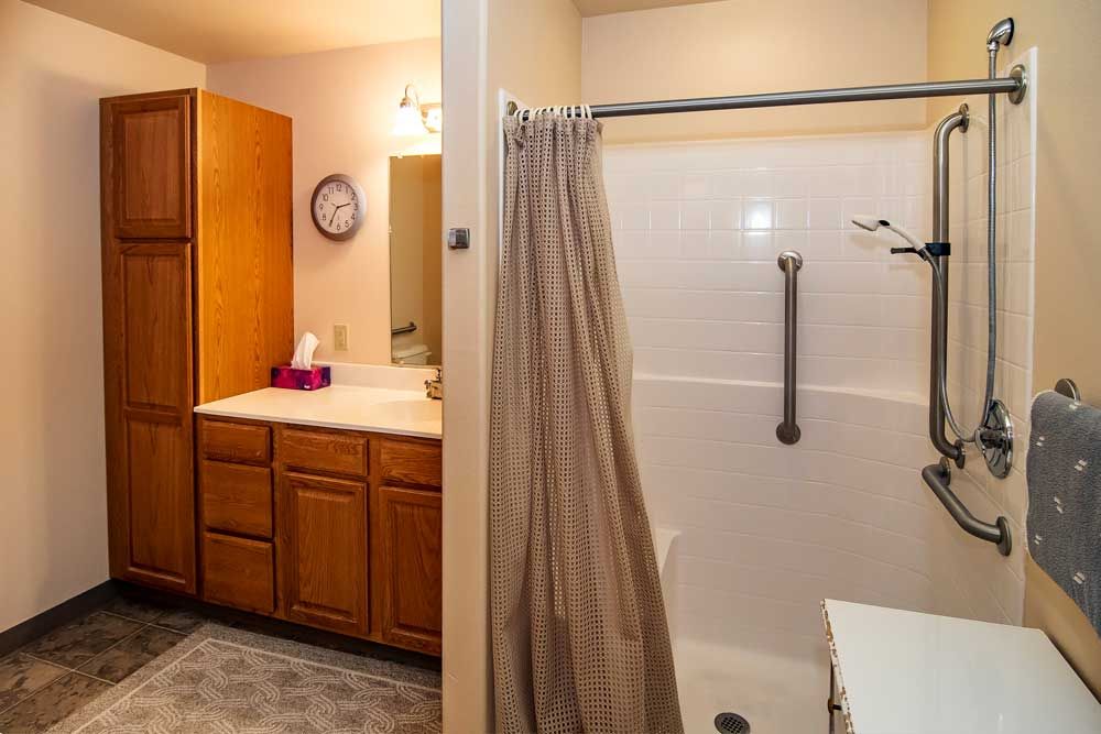 A bathroom with a shower and curtain, featuring a vanity with a sink, mirror, cabinets, and a wall clock.
