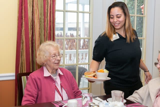A caregiver serves a meal to an elderly woman in a dining setting.