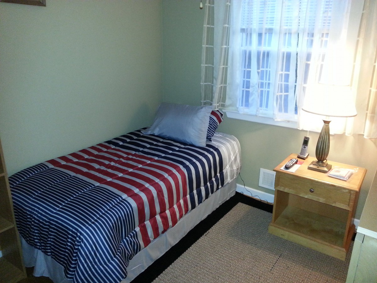 A cozy bedroom featuring a single bed with striped bedding, next to a nightstand with a lamp by the window.