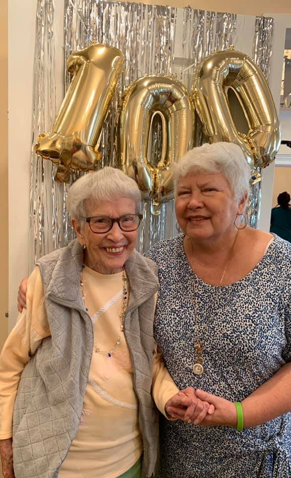 Two elderly women smiling at a birthday celebration with '100' balloons in the background.