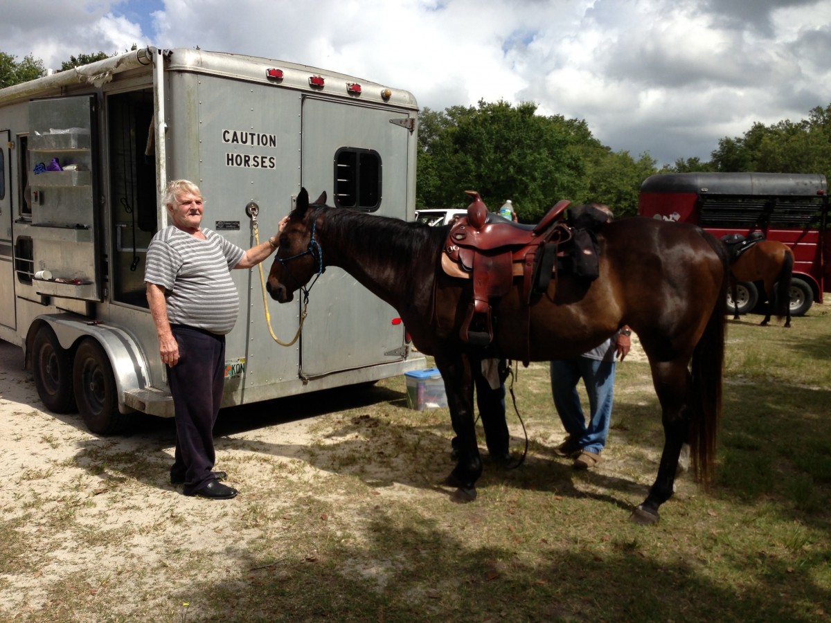 A man stands beside a horse near a trailer in an outdoor setting.