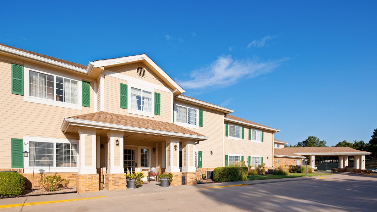 A large two-story residential building with green shutters and beige siding under a clear blue sky.