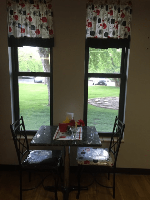 Cozy dining area with a table set for two by large windows overlooking a grassy outdoor scene.