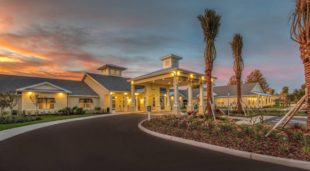 Illuminated building entrance at sunset with landscaped driveway and palm trees.