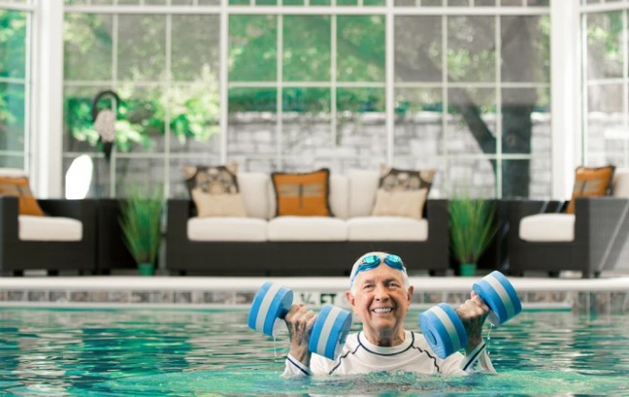 An elderly person enjoys aqua aerobics in an indoor pool, holding water weights and smiling.