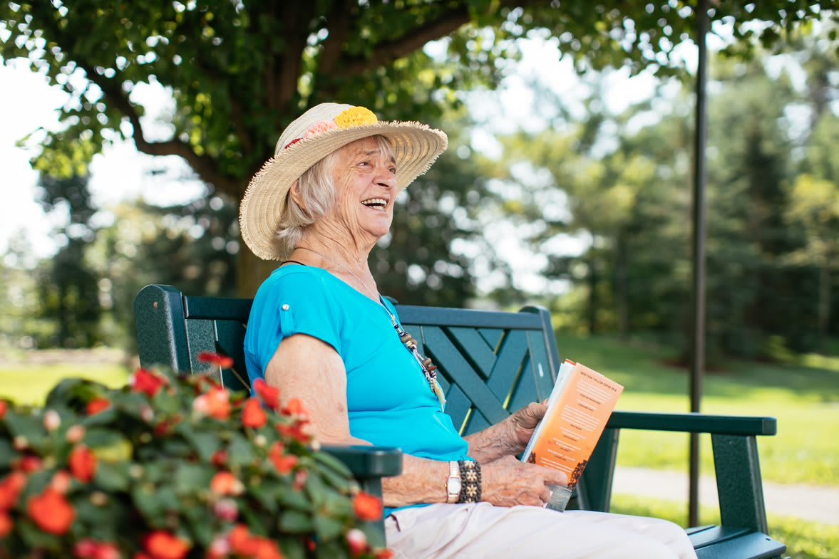 An elderly woman wearing a hat sits on a bench outdoors, enjoying a book.