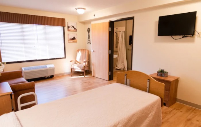 A well-lit hospital room with a single patient bed, armchair, and wall-mounted TV.