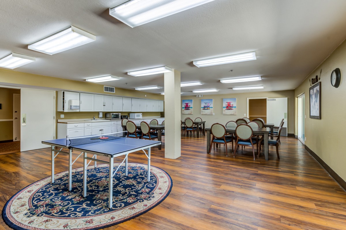 A spacious community recreation room featuring a ping pong table and dining area, illuminated by fluorescent lighting.